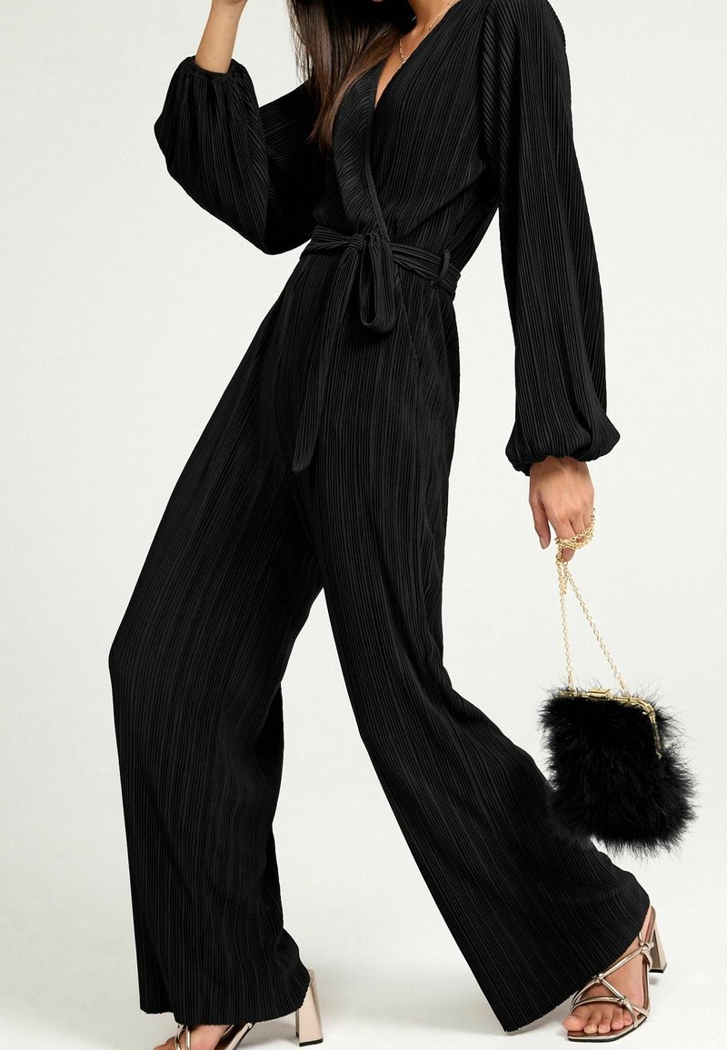 Zwarte geplooide jumpsuit met lange mouwen, wikkelontwerp en tailleband. Gecombineerd met metallic hakken en een kleine zwarte nep-bont handtas.