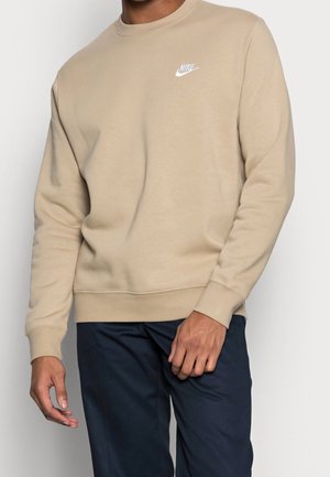 Mężczyzna w beżowej bluzie Nike z białym logo na piersi i ciemnoniebieskich spodniach, stojący na jednolitym tle.