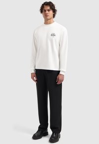 Weißes langärmliges T-Shirt mit rundem Ausschnitt, das ein kleines schwarzes Logo zeigt. Kombiniert mit schwarzen Hosen und schwarzen Schuhen.