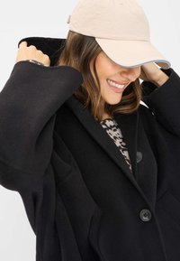 Sort uld-blandede frakke med store ærmer og to knapper, lagd over en mønstret sweater. En beige hat med blå kant fuldender looket.