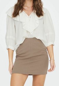 Weiße Bluse mit gerüschtem Kragen und Ärmeln, verziert mit Spitzenakzenten am Ausschnitt, kombiniert mit einem taillierten braunen Miniröckchen.