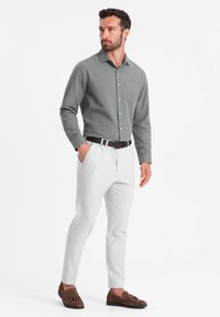 Chemise à manches longues grise pour homme, associée à un pantalon gris clair. La tenue est complétée par des mocassins en daim marron et une ceinture marron.