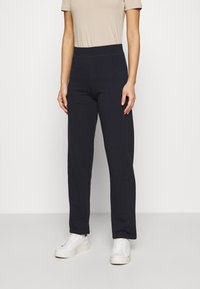 Marina byxor i stretchtyg, med bred midja och rak benpassform, parat med vita sneakers och en beige t-shirt.