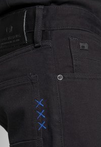 Jeans de denim negro con un acabado texturizado, detalles bordados en azul y botones metálicos. Incluye una etiqueta de cuero en la cintura.