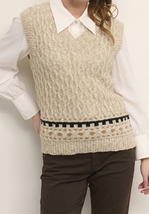 Femme portant un gilet en tricot torsadé beige sans manches avec bordure à motifs sur une chemise blanche à col et un pantalon marron.