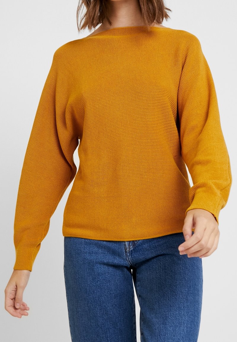 Femme portant un pull en maille jaune moutarde à manches longues associé à un jean bleu, montrée du cou jusqu'aux hanches sur un fond uni.