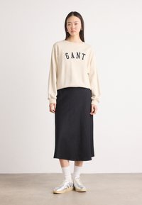 Beige collegepusero, jossa on navy-väriset "GANT"-kirjaimet, yhdistetty mustiin hameeseen, harmaisiin lenkkareihin ja valkoisiin varresta ylöspäin ulottuviin sukkasiin. Yksinkertainen muotoilu ja rento tyyli.