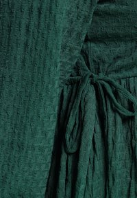 Grön texturerad klänning med ett krinklat mönster, knytmidja och flödande tyg. Materialet verkar lätt och mjukt med intrikata detaljer.