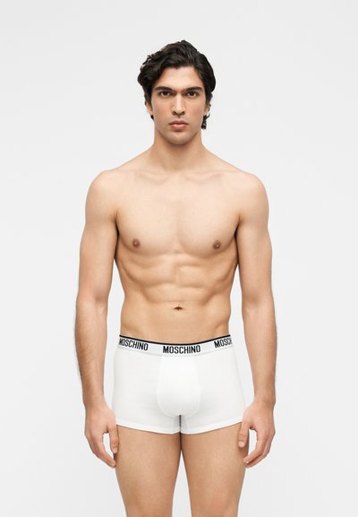 Άνδρας χωρίς πουκάμισο με σκούρα μαλλιά που φοράει λευκά boxer briefs Moschino, στέκεται μπροστά σε μία απλή ανοιχτογκρι επιφάνεια.