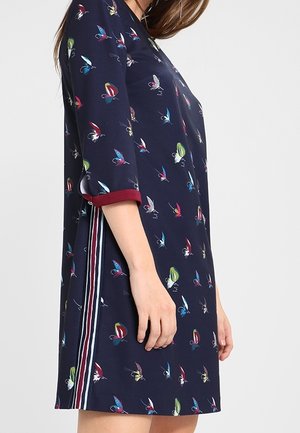 Frau trägt ein marineblaues Kleid mit mehrfarbigen Schmetterlingsmustern, Dreiviertelärmeln und vertikalen roten, weißen und blauen Seitenstreifen.