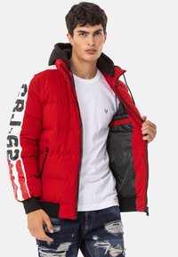 Red puffer jacket with black accents, zip closure, and ribbed cuffs. Features white lettering on the sleeve and a black interior lining.