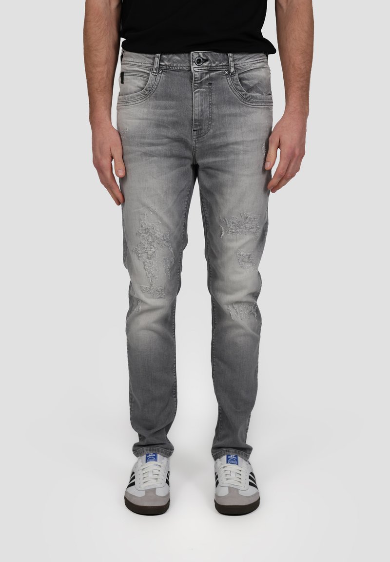 Jean en denim gris avec des patches déchirés, coupe slim, design à cinq poches et fermeture à bouton, accompagnés de baskets blanches.