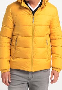 Homme portant une veste matelassée jaune avec des poches zippées et un jean gris, debout les mains détendues le long du corps.