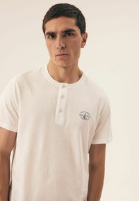 Weißes Baumwoll-T-Shirt mit Rundhalsausschnitt, drei-knopfiger Knopfleiste und blauem gesticktem Logo auf der linken Seite. Glatte Textur, kurze Ärmel.