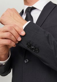 Giacca di suit grigio scuro con una texture liscia, dotata di bottoni neri sui polsini e di un colletto di camicia bianco visibile sotto.