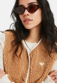 Beige, flauschige Weste mit einer strukturierten Oberfläche und weißen Akzenten, kombiniert mit dunklen Katzenaugen-Sonnenbrillen, getragen über einem hellen Pullover.
