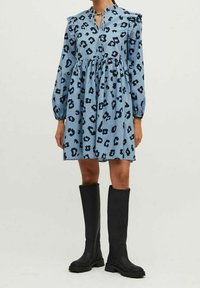 Sininen leopardikuvioinen mekko, jossa on röyhelöillä varustetut olkapäät ja avainaukko kaula-aukko, yhdistettynä korkeisiin mustiin saappaisiin, joissa on teksturoitu pohja.