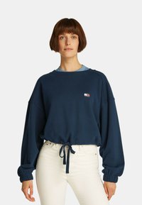 Felpa in cotone blu navy con design cropped, maniche a palloncino e vita legata. Presenta una piccola patch con logo sul petto.