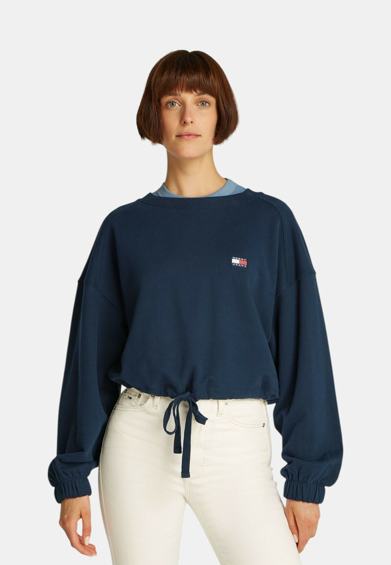 Felpa in cotone blu navy con design cropped, maniche a palloncino e vita legata. Presenta una piccola patch con logo sul petto.