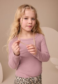 Bluză mov deschis cu mâneci lungi, cu dungi, asortată cu pantaloni bej cu imprimeu leopard. Copilul ține o agrafă de păr, având părul blond, lung și ondulat, cu împletituri.