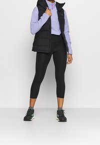 Kobieta ubrana w czarne krótkie legginsy, czarną kamizelkę puchową, fioletową bluzę z półsuwnikiem, szare skarpetki oraz czarne buty sportowe z zielonymi akcentami.