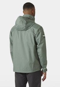 Chaqueta resistente al agua de color verde claro, con capucha, cierre de cremallera y logo en la manga. Presenta una textura suave y un corte ajustado.