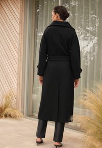 Long manteau noir en laine avec une ceinture, un large col et des poignets retroussés, porté sur un pantalon en cuir noir. Texture lisse, design minimaliste.