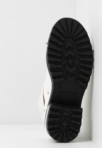 Baskets blanches avec une tige texturée et une semelle épaisse en caoutchouc noir. Présentent un motif de crampons proéminent pour l'adhérence et des accents métalliques argentés.