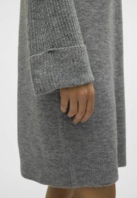 Abito grigio lavorato a maglia con una superficie texture, maniche ampie e polsini a costine. Il tessuto appare morbido e caldo, ideale per il clima freddo.