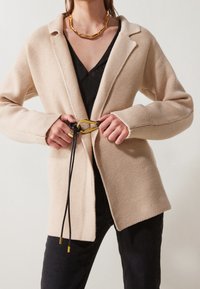 Beige gestrickte Strickjacke mit übergroßen Revers, einer Bindeschließe mit einem goldenen Akzent und langen Ärmeln; getragen über einem schwarzen Oberteil und einer Hose.