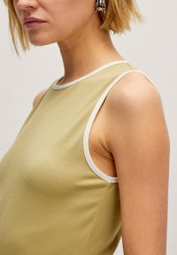 Mouwloze jurk van lichtgroene stof met een gladde textuur, met witte afwerking langs de halslijn en armsgaten.