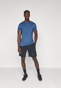 Blå atletisk t-skjorte med korte ermer, med en svart logo på venstre erme, kombinert med svarte shorts som viser den samme logoen.