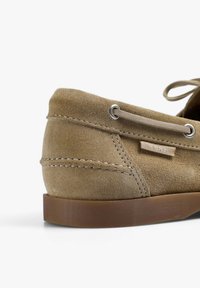 Dettaglio della parte posteriore e laterale di un mocassino in suede beige con una suola marrone e un laccio in pelle infilato attraverso occhielli di metallo.