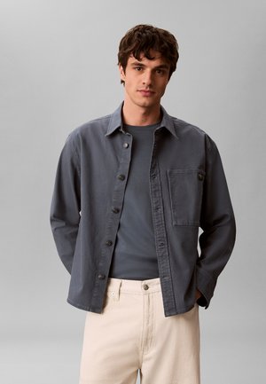 Ung mand iført en mørkegrå skjorte med knapper oven på en matchende t-shirt og lysebeige bukser, stående mod en ensfarvet grå baggrund.