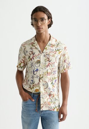 Giovane uomo con occhiali e camicia a maniche corte con bottoni decorata con motivi colorati di stelle e lune, abbinata a jeans blu.