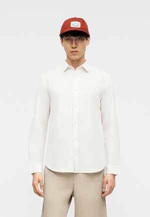 Man in beige broek en wit overhemd met rode pet met wit rechthoekig logo, staand tegen een effen lichtgrijze achtergrond.