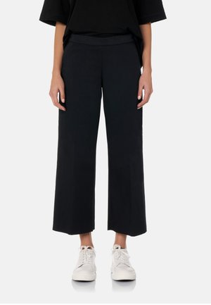 Personne portant un pantalon noir large et court ainsi que des baskets blanches, debout devant un fond clair uni.