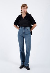 Höga blå denimjeans med smalnande ben, minimal sömnad och klassisk knappstängning, i kombination med en svart ribbad topp och en kortärmad kofta.