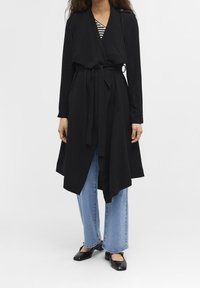 Schwarzer langer Mantel mit drapiertem Vorderteil, in der Taille gebunden, kombiniert mit blauen Weitbeinjeans und schwarzen Schuhen, vor einem schlichten Hintergrund.