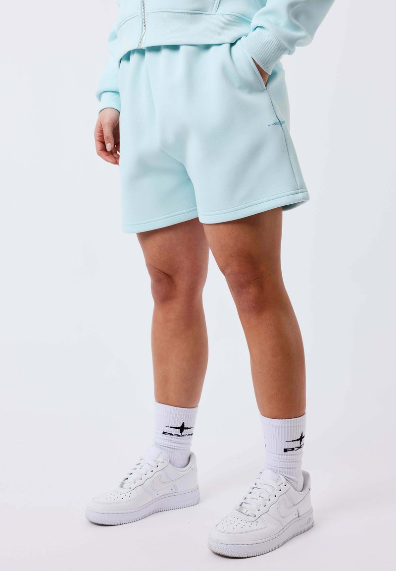 Personne portant un short bleu clair et un sweat à capuche assorti, des chaussettes blanches de marque montantes et des baskets blanches Nike, debout les mains dans les poches.
