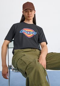 Grå bomull t-skjorte med et fargerikt "Pickies"-grafikk, parret med olivengrønne cargo-bukser. Modell sitter på en klar stol mot en ensfarget bakgrunn.