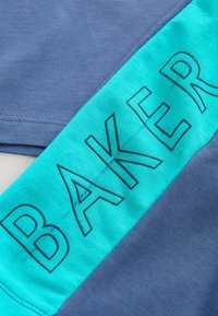 Zila T-kreklu audums ar gludu tekstūru; tam ir drosmīga tirkīza lente ar melnu uzrakstu "BAKER". Dizains ir vienkāršs un mūsdienīgs.