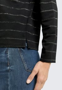 Schwarzes Oberteil mit silbernen horizontalen Streifen, strukturierter Stoff, lange Ärmel und kleiner seitlicher Schlitz am Saum, kombiniert mit blauem Denim.