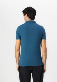 Teal polo skjorte med klassisk krage, korte ermer og en glatt tekstur. Bært med svarte jeans, som viser minimalt med detaljer bak.