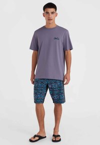Camiseta de algodón morada con un pequeño logo y pantalones cortos de baño negros que presentan un patrón geométrico en azul y rosa. Las chanclas negras completan el look.