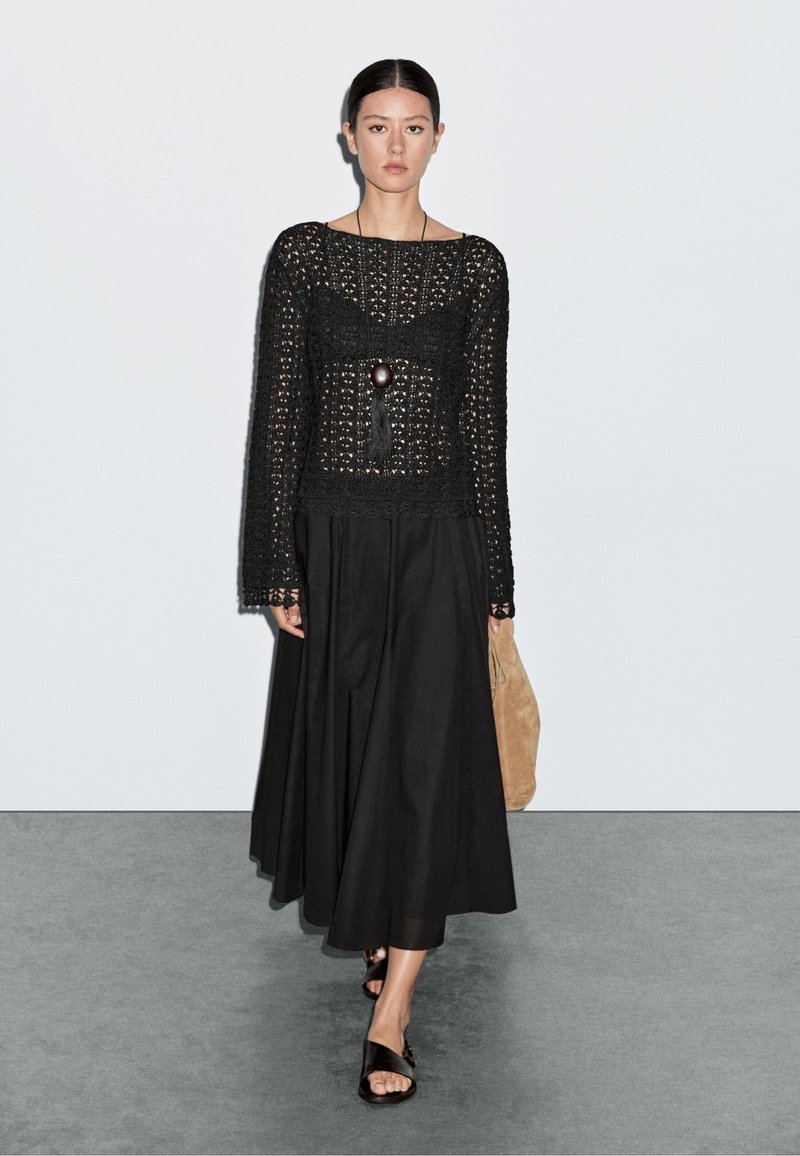 Top in crochet nero con maniche lunghe, abbinato a una gonna nera. Include un ciondolo con nappina e una borsa color cuoio. Sandali neri completano il look.