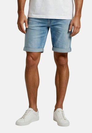 Man wearing light blue rolled-up denim shorts, white sneakers, and a white shirt standing against a plain background.