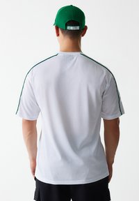 Camisa atlética blanca de malla con detalles verdes en los hombros, mangas cortas y cuello redondo. Acompañada de una gorra de béisbol verde.