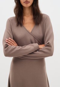 Hnědé zavinovací šaty s dlouhými rukávy, jemnou texturou a hlubokým výstřihem do V. Mají úzkou korzetovou část a splývavou sukni. Minimalistický design.