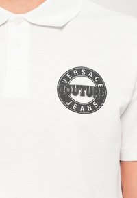 Λευκό polo μπλουζάκι με μαύρο κυκλικό λογότυπο Versace Jeans Couture στο αριστερό στήθος, διαθέτοντας γιακά με κουμπιά και κοντά μανίκια.